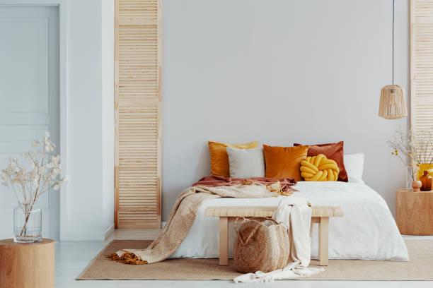 Brown and orange pillows on white bed in natural bedroom interior with wicker lamp and wooden bedside table with vase