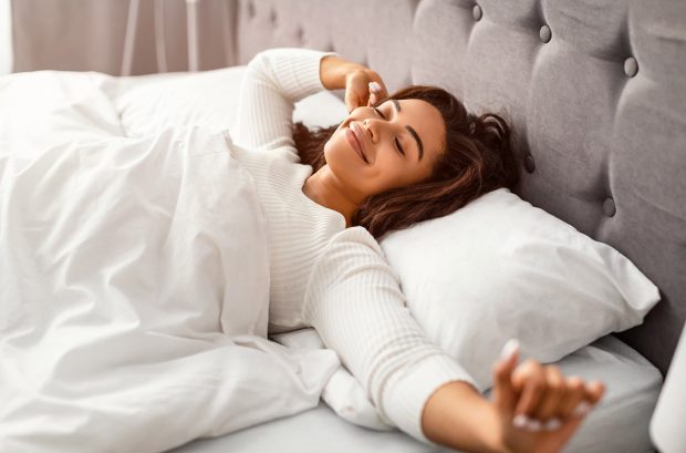 Rest And Relax Concept. Beautiful peaceful black lady stretching arms after wakeup, enjoying sunny morning, millennial African American female lying under blanket in bed with white bedsheets