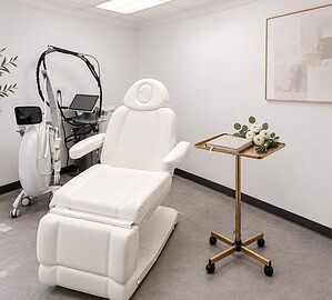 Editorial illustration inspired by the practice’s treatment room, with a planner and flowers. Editorial illustration of a bright white treatment room with a white reclining chair and a slim gold rolling tray holding a closed linen planner and a small bunch of white ranunculus with eucalyptus
