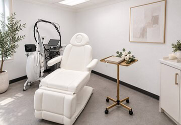 Editorial illustration of a bright white treatment room with a white reclining chair and a slim gold rolling tray holding a closed linen planner and a small bunch of white ranunculus with eucalyptus